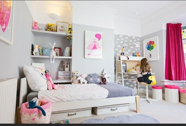 Bright, colorful girls bedroom with white furniture, pink accents, floating shelves, wall art, and storage drawers under the bed